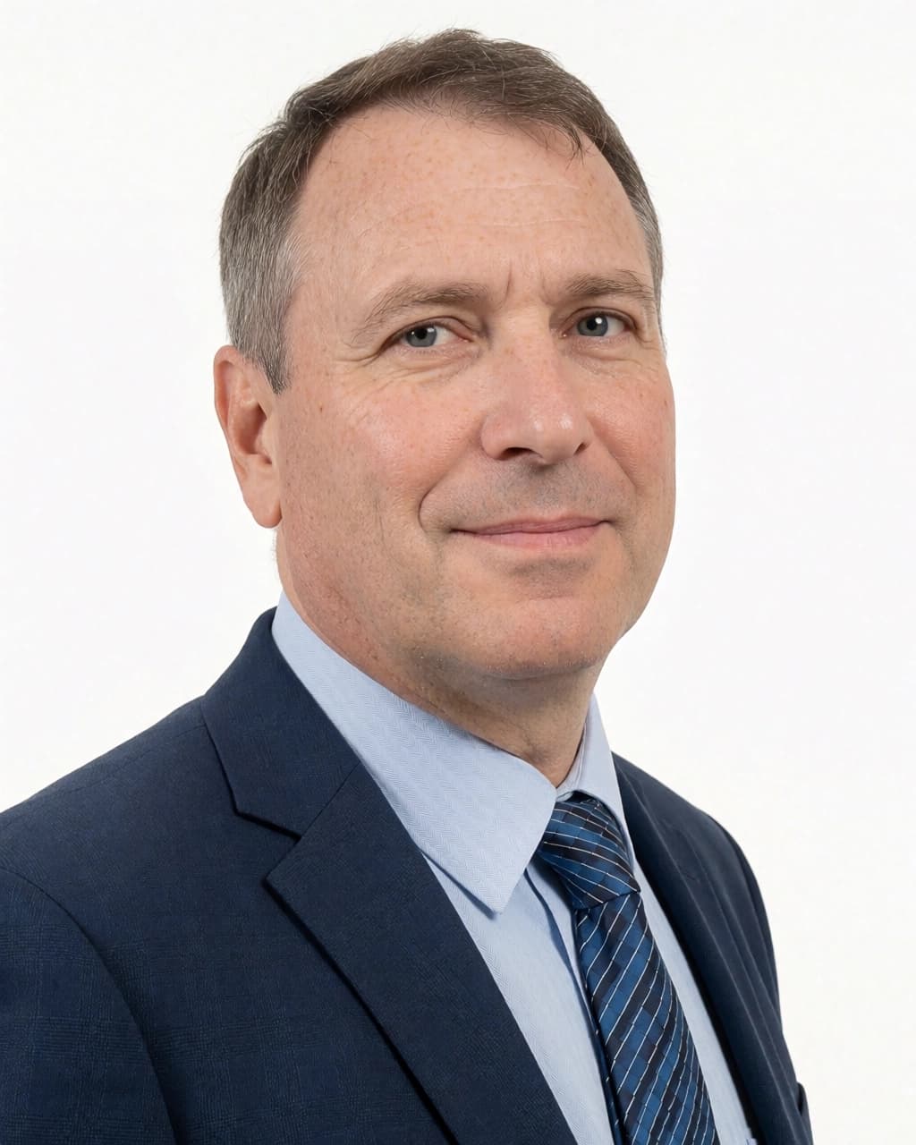 A man with short brown hair and light skin is shown in a head-and-shoulders studio portrait against a white background. He is wearing a dark blue suit jacket, a light blue dress shirt, and a blue and black striped tie. He is looking directly at the camera with a subtle, closed-mouth smile.