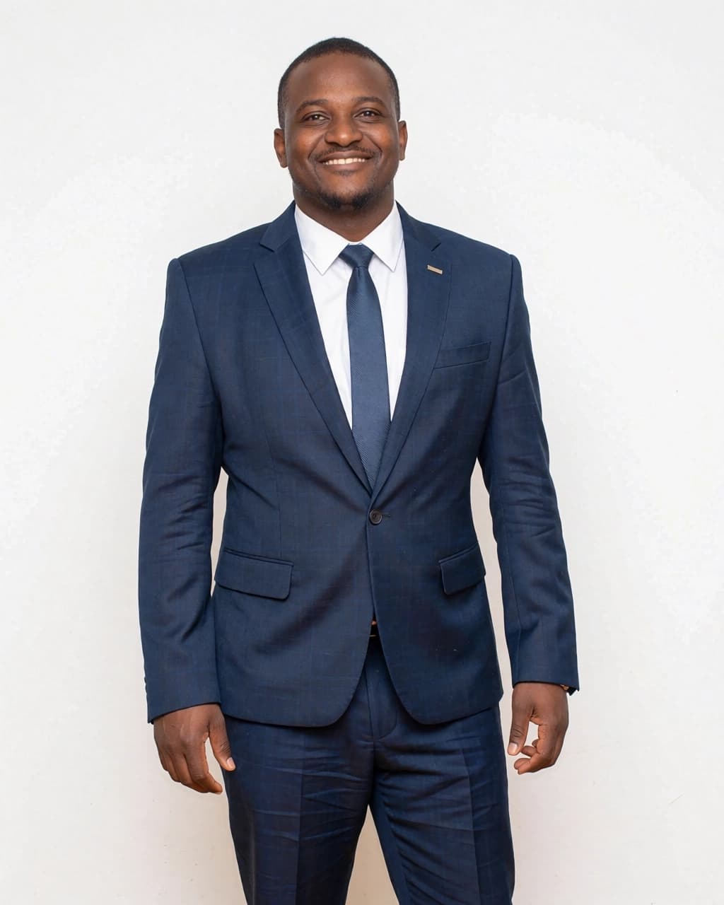 A man with dark skin and short hair is wearing a navy blue suit, a crisp white dress shirt, and a matching navy blue tie. He is standing against a plain white background and is smiling directly at the camera with his mouth slightly open. He has a small, light-colored lapel pin on his left collar. His hands hang relaxed at his sides.