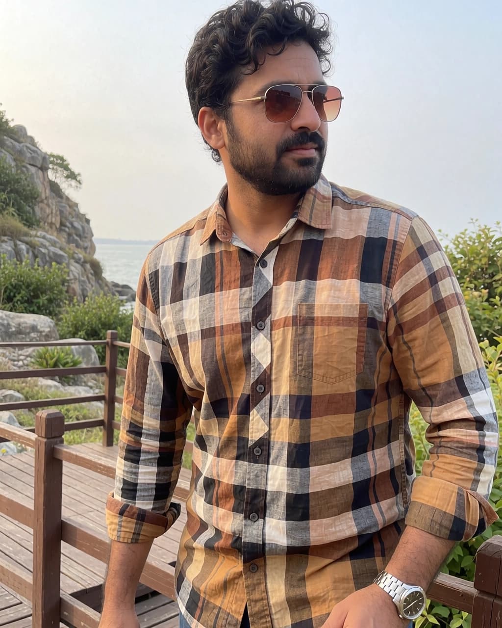 A man with dark, wavy hair and a beard stands outdoors while looking to his right. He is wearing aviator-style sunglasses and a button-down shirt with a brown, black, and white plaid pattern. His sleeves are rolled up to his elbows, revealing a silver wristwatch on his left wrist. He is positioned on a wooden deck with railings, set against a backdrop of rocky cliffs, greenery, and a body of water in the distance under a light sky.