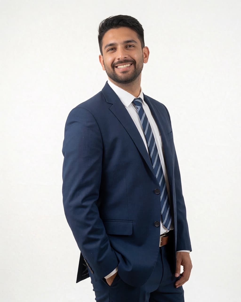 A young man with dark hair and a short beard stands against a plain white background. He is wearing a dark blue suit jacket, a white dress shirt, and a blue tie with diagonal white stripes. He is facing toward the left while looking directly at the camera with a smile. His right hand is placed inside his jacket pocket.