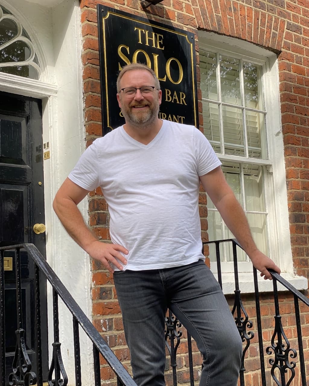 A middle-aged man with a beard and glasses stands on the steps in front of a brick building. He is wearing a white V-neck t-shirt and grey jeans. His right hand is on his hip, and his left hand rests on a black metal railing. Behind him, a black sign with gold lettering reads "THE SOLO BAR & RESTAURANT." To the left is a black door with a gold handle, and to the right is a white-framed window.