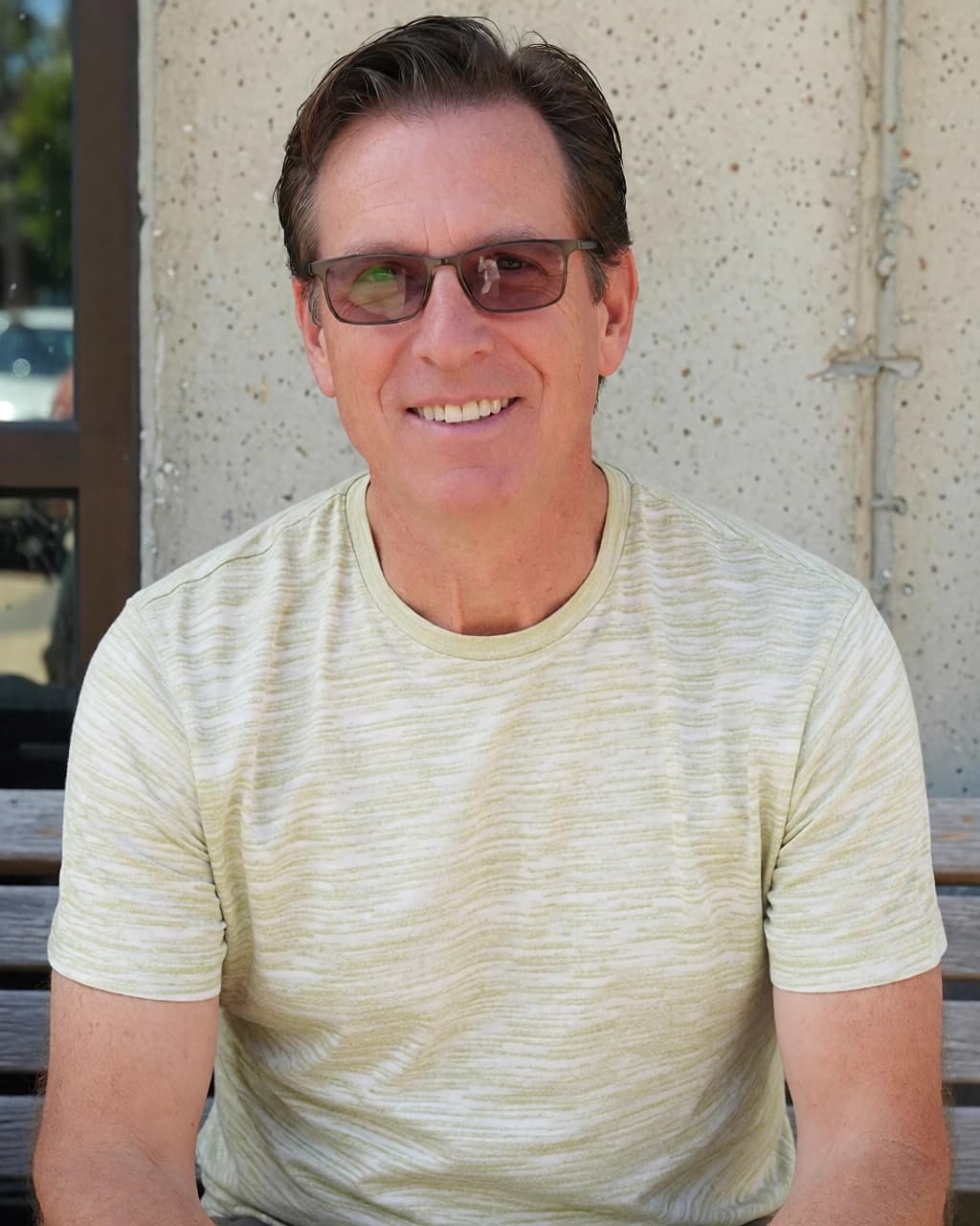 A middle-aged man with short brown hair and eyeglasses is smiling directly at the camera. He is wearing a light green, horizontally-textured short-sleeved t-shirt. The man is sitting on a wooden bench in front of a light-colored, textured concrete wall. To his left, there is a dark-framed window or door with a reflection visible in the glass.