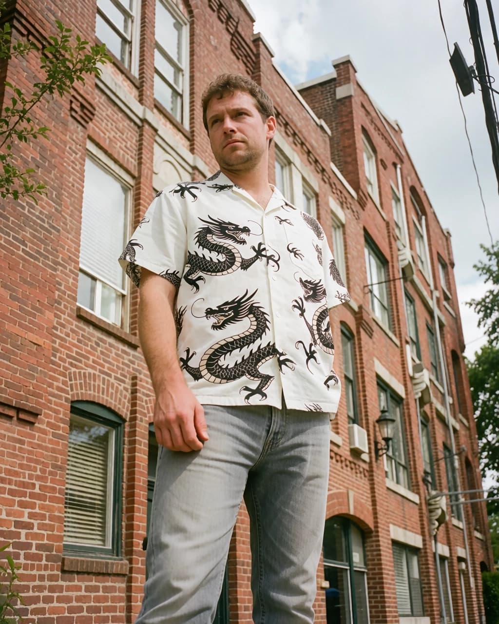 A light-skinned man with short brown hair stands outdoors, viewed from a low angle. He is wearing a short-sleeved, white, button-down shirt featuring a black dragon print and light grey denim jeans. He stands in front of a multi-story red brick building with multiple rectangular windows. A portion of a green tree is visible on the left side of the frame, and a dark utility cable hangs in the upper right corner against a pale sky.