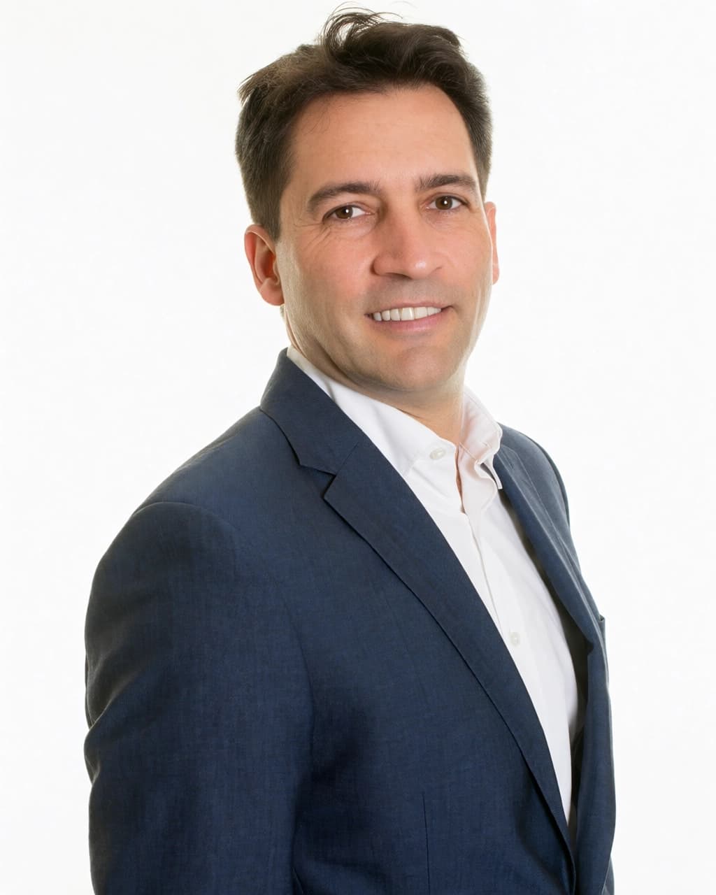 A man with short brown hair and brown eyes is smiling at the camera. He is wearing a white collared shirt under a navy blue blazer. The background is a plain white surface. The man is positioned in profile, looking towards the right side of the frame.