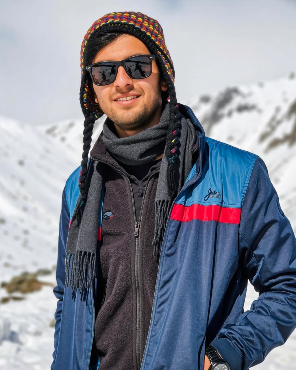 A smiling man is wearing a multi-colored knit hat with braided tassels and black sunglasses. He is dressed in a dark grey scarf, a black fleece jacket, and a blue jacket with a red horizontal stripe across the chest. His left hand is inside his jacket pocket, revealing a black watch on his wrist. The background features snow-covered mountains under a bright sky.