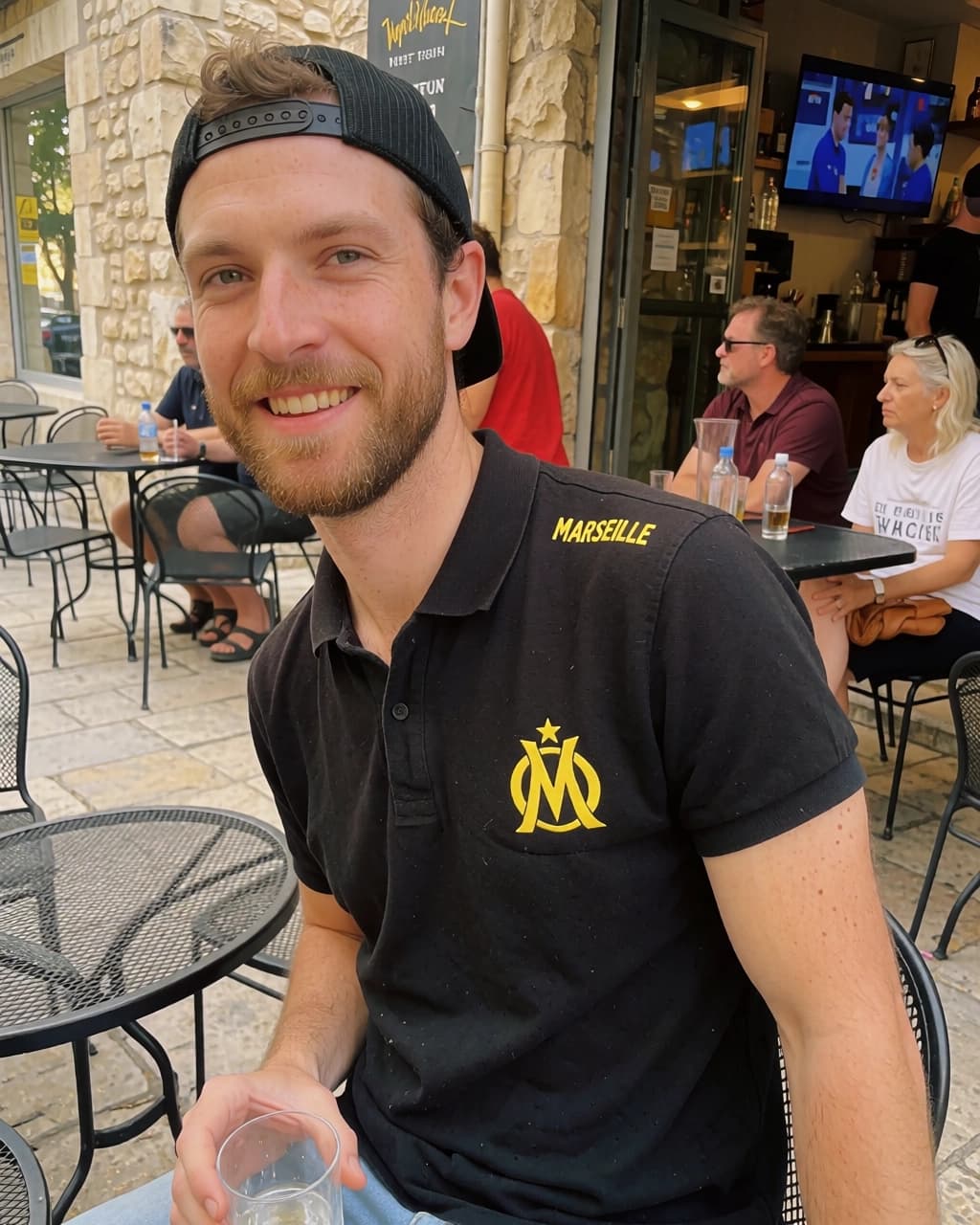 A man with a brown beard, wearing a black cap and a black polo shirt with yellow text and a logo, smiles at the camera while sitting at a metal table outdoors. He holds a small clear plastic cup. In the background, other people are seated at tables on a stone patio, and a television is mounted inside an open doorway.