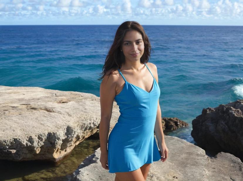 wide portrait photo of a latina woman with dark hair standing on a beach rock, wearing a light blue mini dress, ocean in the background