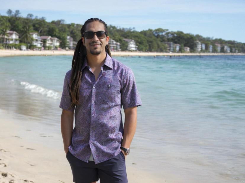 wide portrait photo of a latino man with dreadlocks standing casually on a beach, wearing a paisley shirt, shorts, and sunglasses, sea shore in the background
