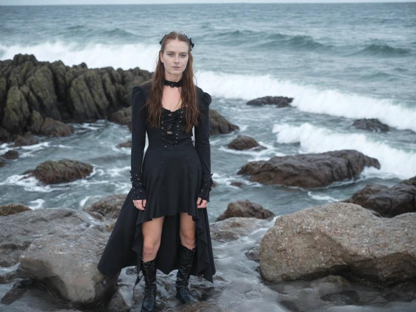 wide portrait photo of a caucasian woman with auburn hair standing on a rocky beach shore, wearing a black high-low gothic sleeved dress and boots, wavy sea in the background