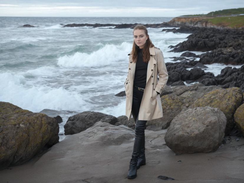 wide portrait photo of a caucasian woman with auburn hair posing on a rocky beach, wearing a beige trench coat over a black shirt, black pants, and black boots, furious sea in the background