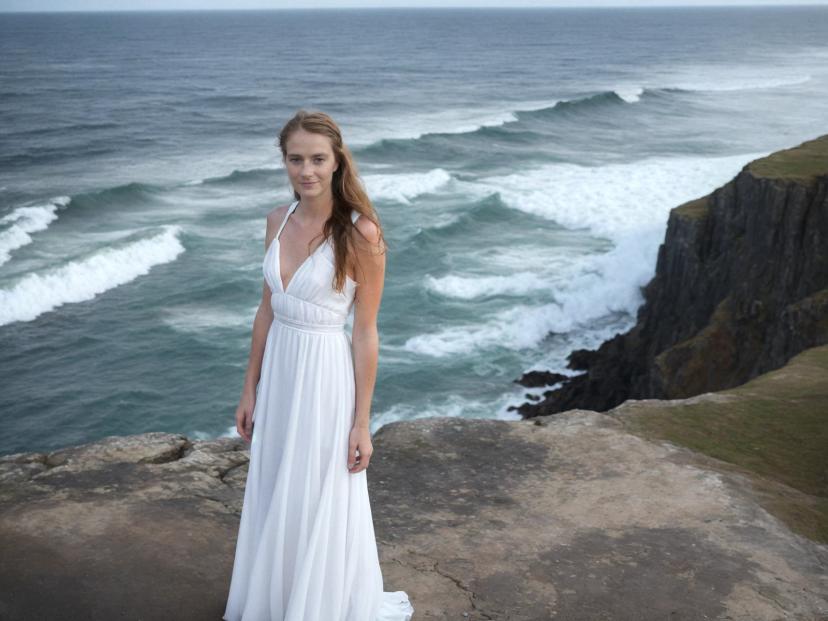 wide portrait photo of a caucasian woman with strawberry blonde hair standing on a sea cliff, wearing a beautiful flowing white dress, wavy sea in the background