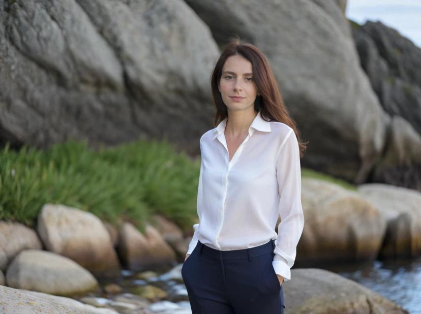 wide portrait photo of a beautiful caucasian woman with auburn hair standing on a rocky beach, wearing a buttoned white shirt and navy trousers, rock formations on the background