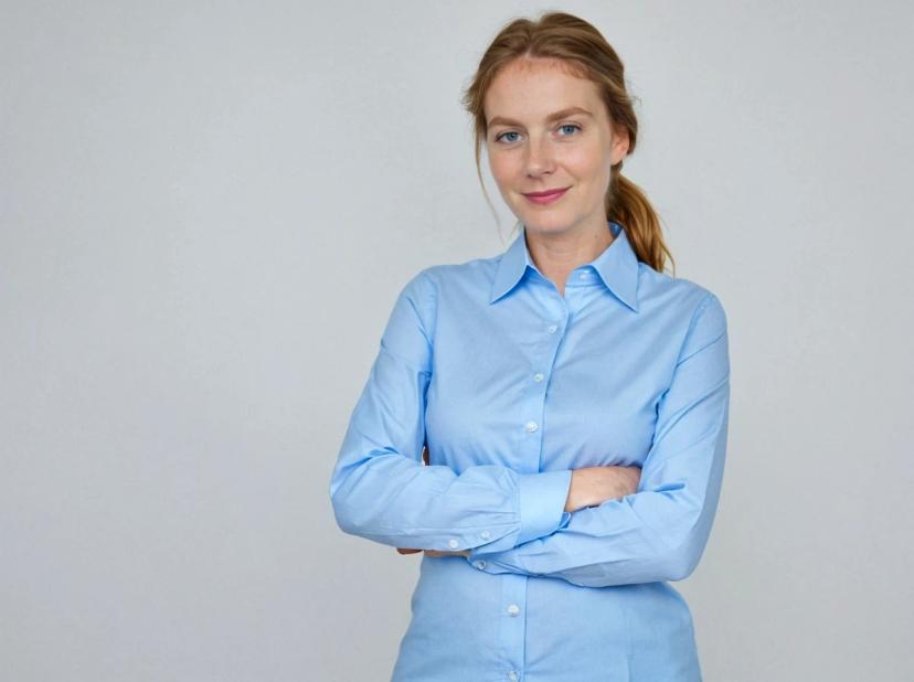 professional headshot of a young woman with tied hair and her arms crossed standing against a solid white background. She is wearing a light blue button-up shirt