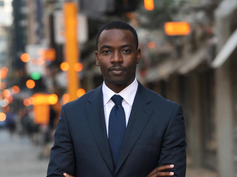 professioanl headshot of a young black man with short hair and his arms crossed standing with a confident expression in a city with lights in the background. He is wearing a navy suit and a blue tie