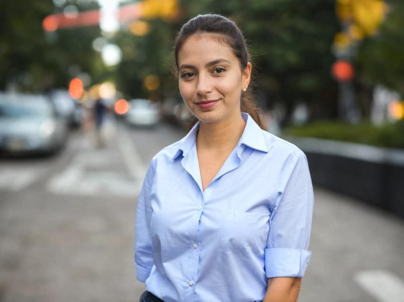 professioanl headshot of a pretty business woman with a slight smile, standing in a modern city street with cars in the background. She is wearing a light-blue button-up shirt