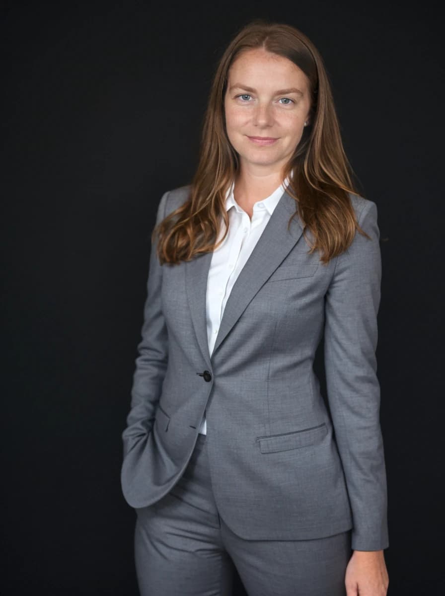 professional business photo of a caucasian woman with ginger hair and a smile stading against a solid black background. She is wearing a dark gray business pantsuit and a white shirt