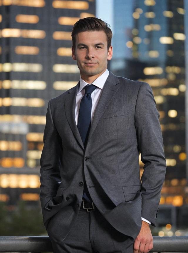 Professional business photo of a young man in a charcoal suit and white shirt with a blue tie standing outdoors with illuminated modern office buildings in the background