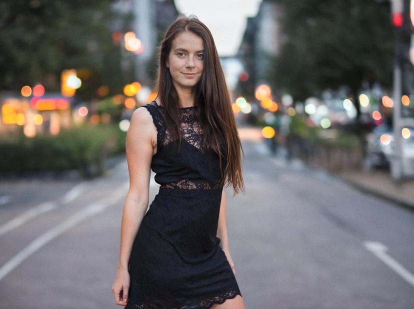 A woman wearing a black lace dress is standing in the middle of a street with backdrop of city lights and traffic at twilight.