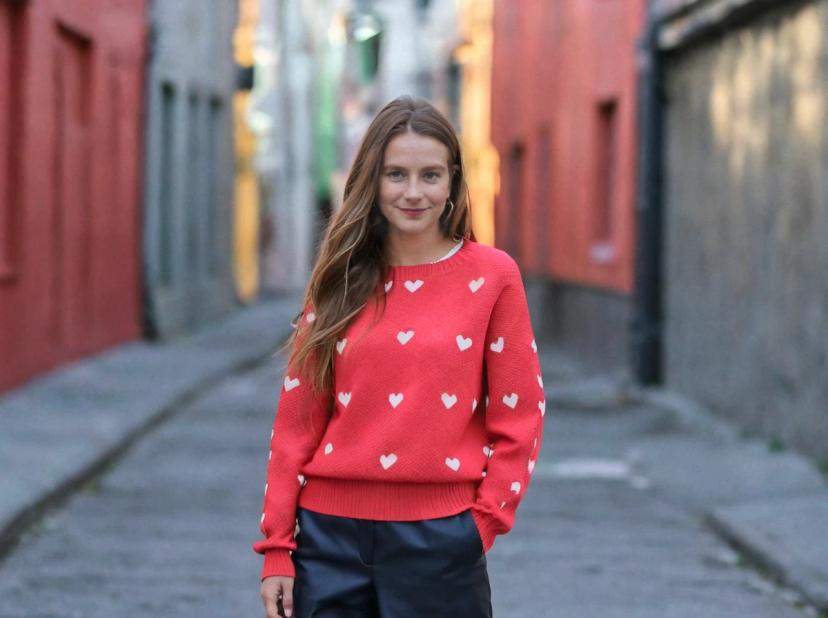 A woman standing in an alleyway wearing a red sweater with white hearts and black pants. There are colorful buildings in the background with urban scenery.