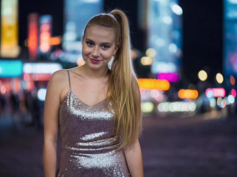 A woman with long blond hair, wearing a sparkling sleeveless dress, standing in front of a vibrant cityscape at night with illuminated billboards and colorful city lights.