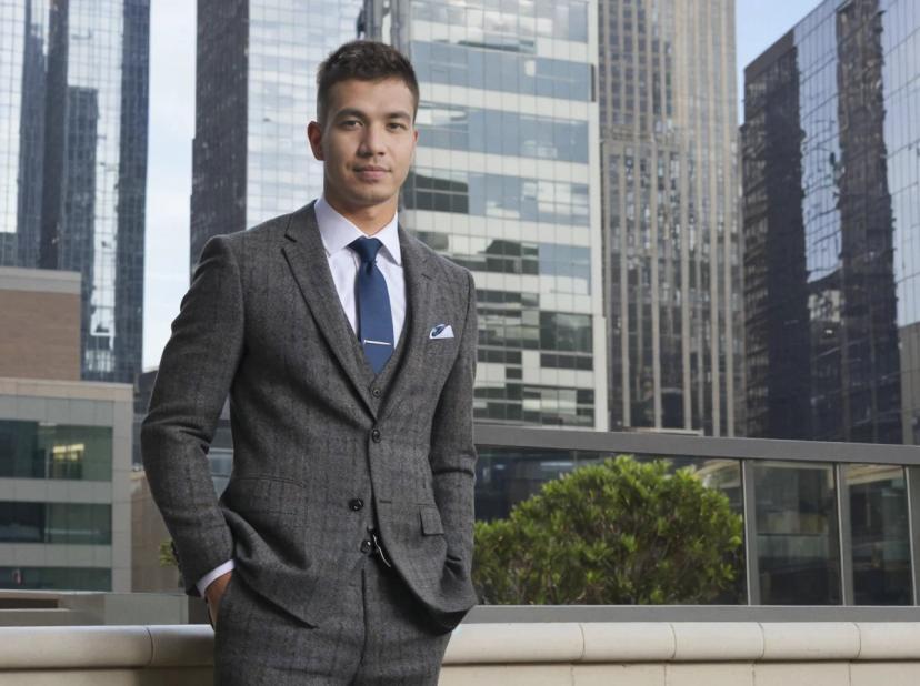 A man in a tailored grey suit with a blue tie and pocket square standing in front of skyscrapers.