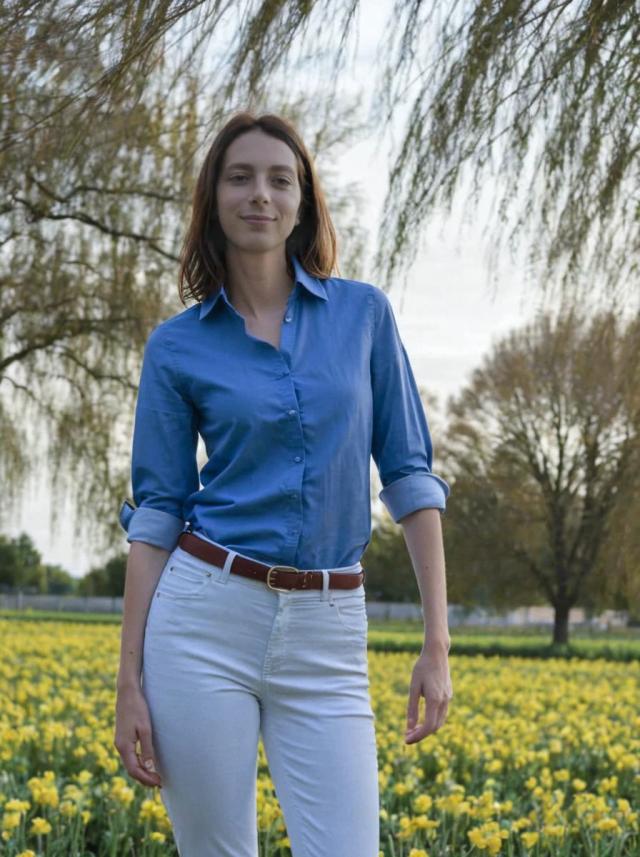 portrait photo of a beautiful woman with a shy smile standing on a beautiful yellow flower field with trees, she is wearing a blue buttoned shirt and white trousers with a belt
