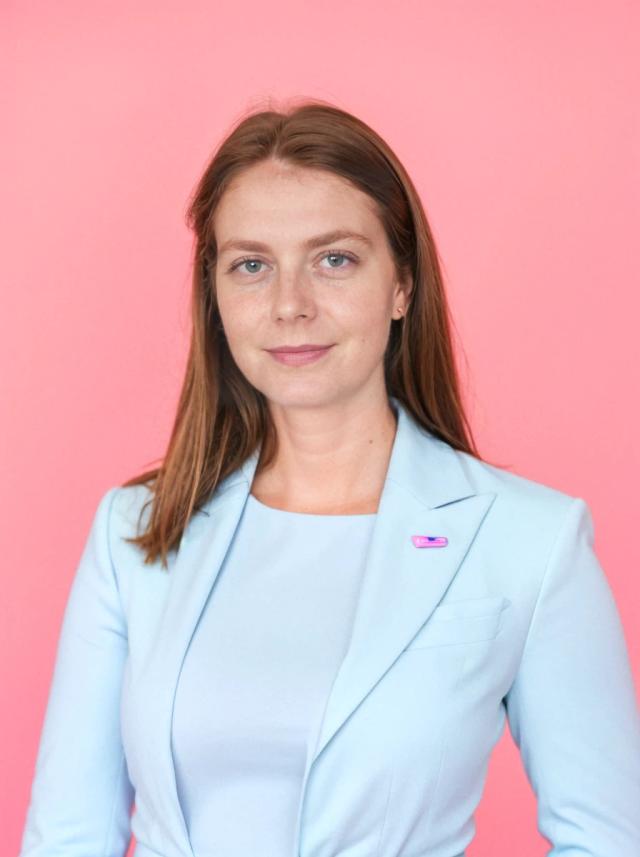 professional headshot of a young woman with a confident expression and straight hair posing against a solid pink background. She is wearing a matching light-colored blazer and shirt 