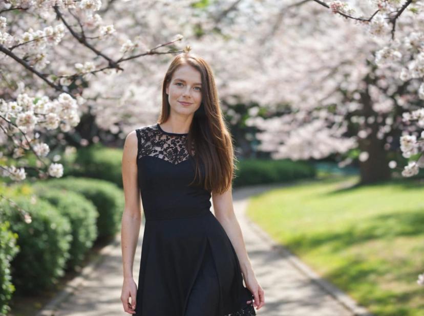A woman in a black sleeveless dress with lace detailing walks along a path surrounded by blooming cherry blossom trees in a lush garden.