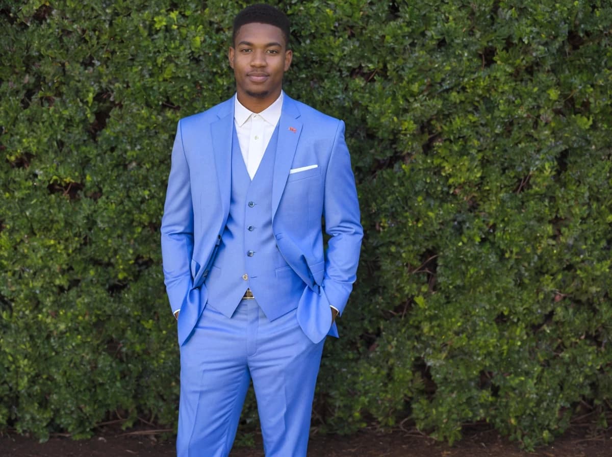 A man dressed in a stylish bright blue three-piece suit with a white shirt and a pocket square stands confidently in front of a dense green hedge.