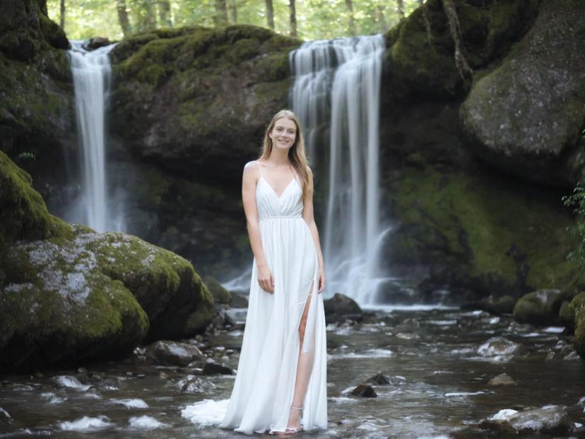 A woman in a white dress standing in front of a tranquil waterfall surrounded by lush greenery. Rocks covered with moss line the stream, and the water cascades gracefully down multiple levels into a serene pool below.