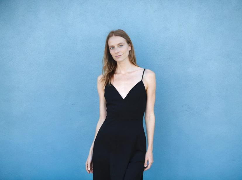 A woman in a black sleeveless jumpsuit standing against a blue textured wall.