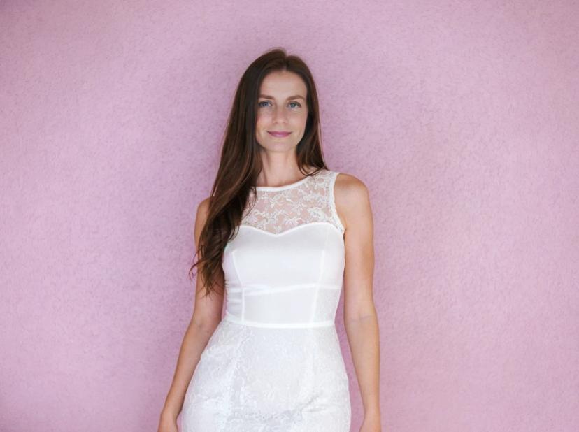 A woman wearing a white lace wedding dress, standing against a pink textured background. She has long, dark brown hair.