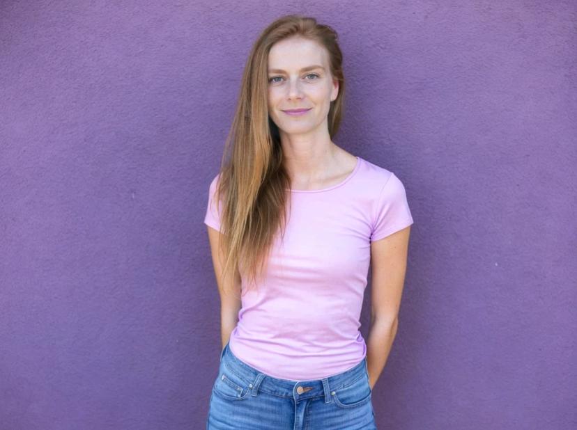 A woman wearing a pink t-shirt and blue denim jeans stands against a purple wall.