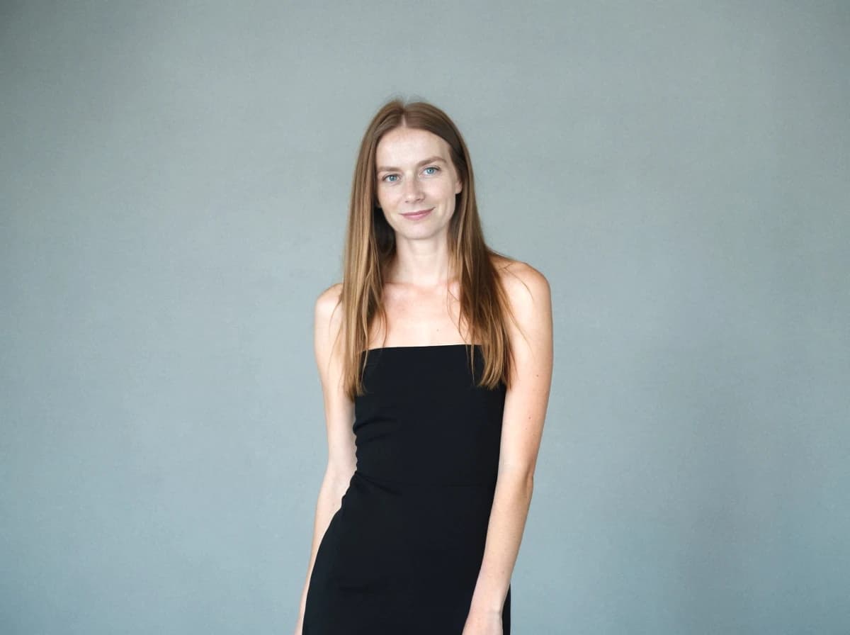 A woman with long blonde hair wearing a black sleeveless dress stands against a gray background.