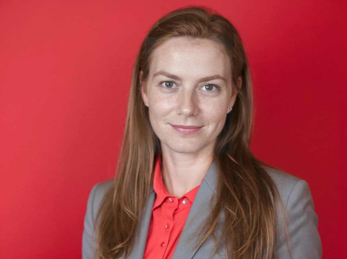 A woman with long hair wearing a gray blazer and red blouse stands in front of a solid red background.