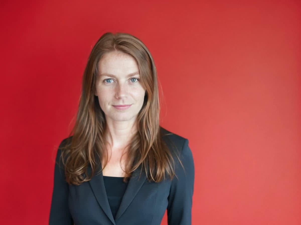 A woman with long brown hair wearing a dark blazer stands in front of a solid red background.