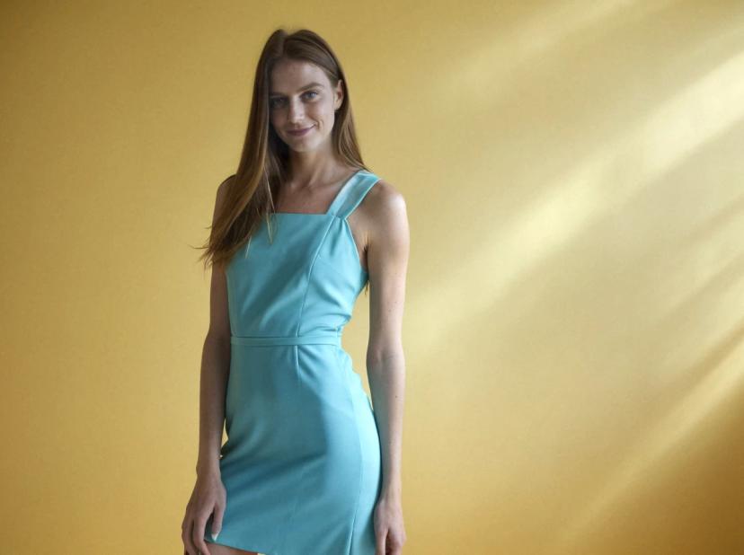 A woman with long hair wearing a pastel blue dress stands against a yellow background with soft light rays casting down. She is facing sideways, showcasing the profile view of the outfit.