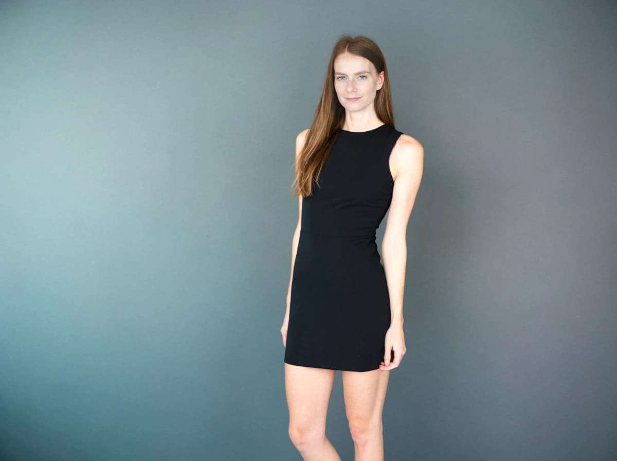 A woman standing against a blue-gray background, wearing a sleeveless black mini dress with a fitted silhouette, their hands resting gently by their sides.