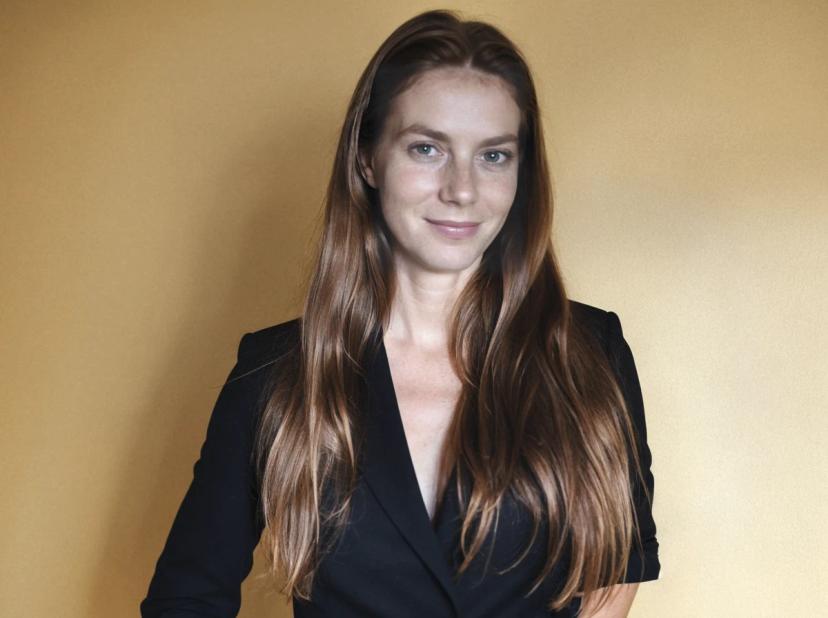 A woman with long brown hair wearing a black blazer standing against a yellowish wall.