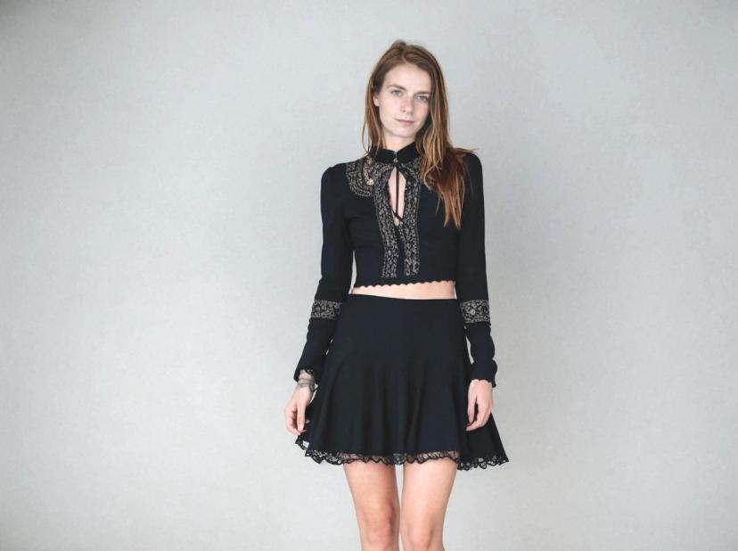 A woman wearing a black long-sleeve crop top with lace detailing and a matching black skater skirt, standing against a grey background.