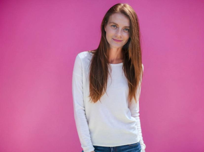 A woman with long brown hair wearing a white long-sleeved shirt and blue jeans standing against a bright pink background.