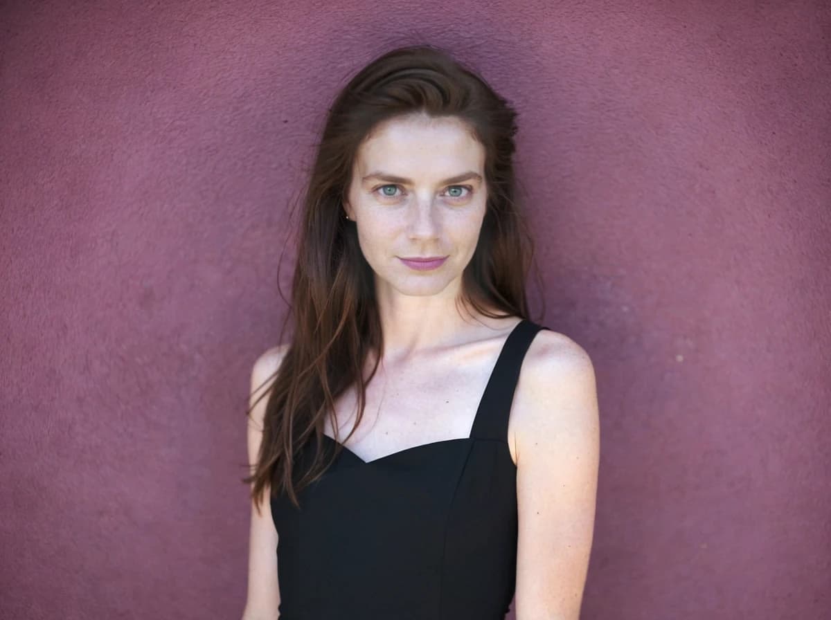 A woman with long brown hair wearing a black sleeveless dress stands against a textured purple wall.