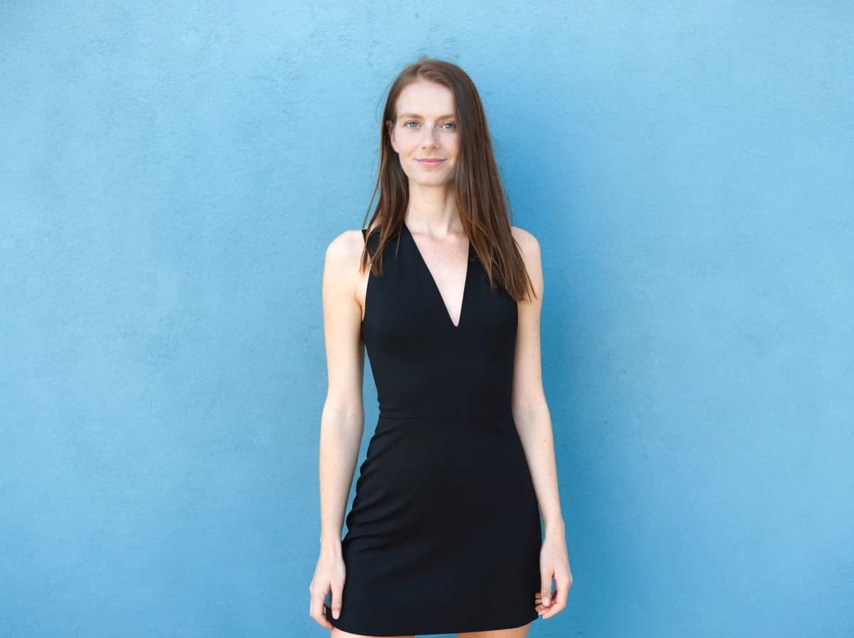 A woman wearing a sleeveless black dress standing in front of a plain blue wall.