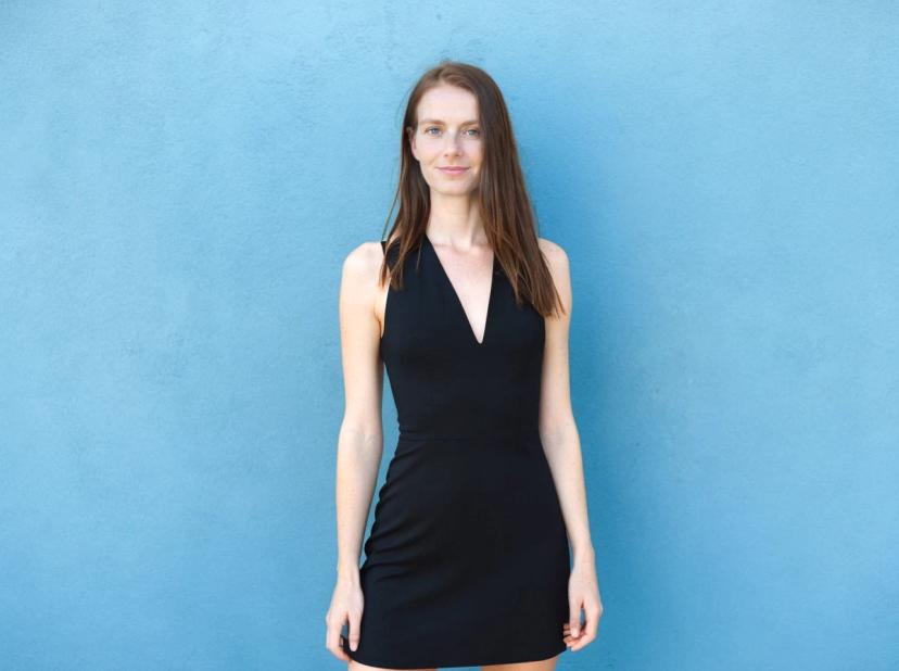A woman wearing a sleeveless black dress standing in front of a plain blue wall.