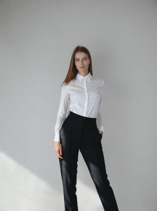 A woman stands against a grey background, dressed in a white button-up shirt and black high-waisted trousers with hands slightly at their sides.