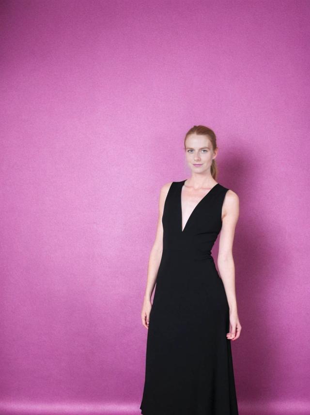 A woman in a black sleeveless v-neck gown standing against a pink textured background.