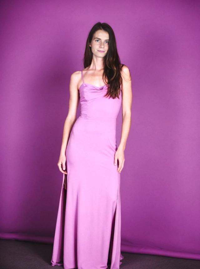 A woman standing against a purple background wearing a sleeveless, floor-length lavender dress with a draped neckline and thin straps.