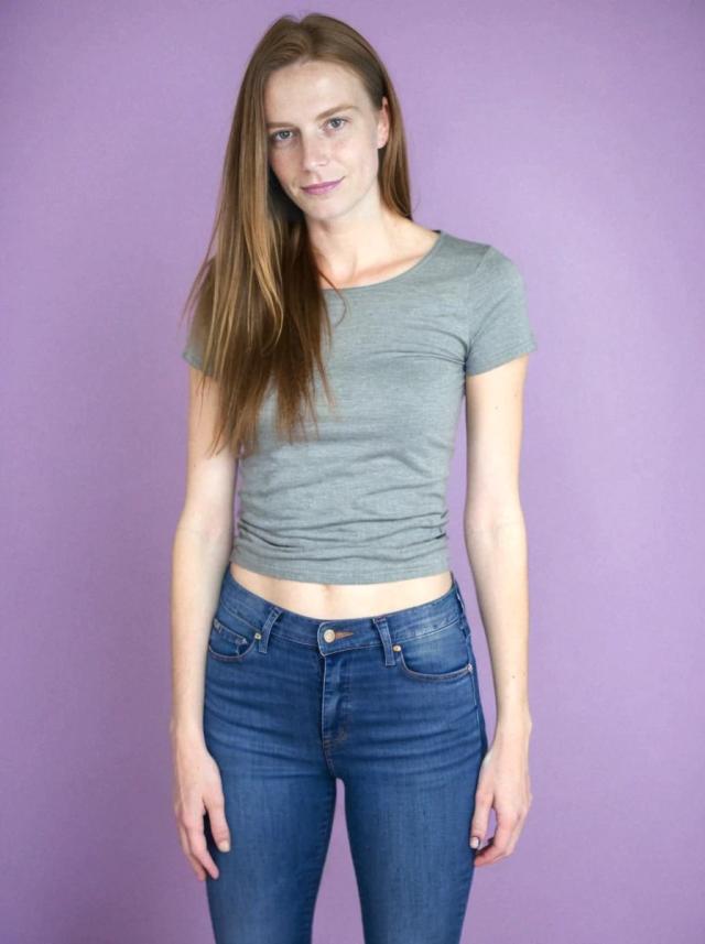A woman wearing a gray crop top and blue jeans standing against a purple background.