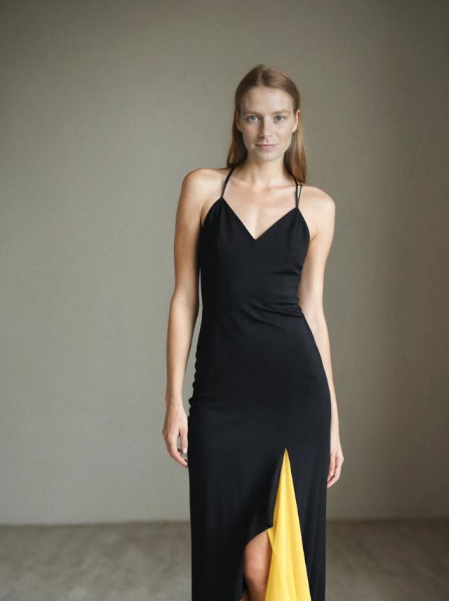 A woman standing in a minimalist setting wearing a sleek black dress with spaghetti straps and a high slit revealing a yellow garment underneath. The background is a plain, light grey wall, and the flooring is a neutral shade.