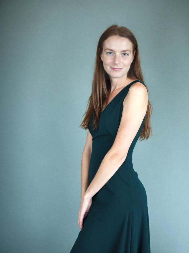 A woman wearing a dark green sleeveless dress stands against a solid grey background, with their body angled to the side and one arm resting along their body.